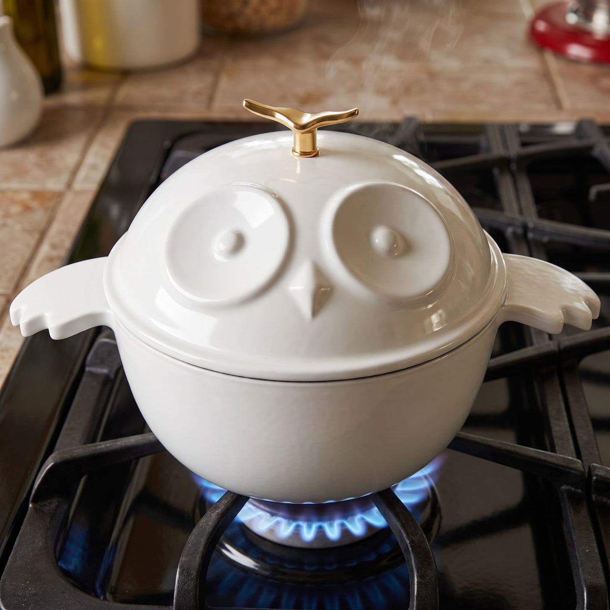 A white cast iron owl-shaped pot with a matching lid sits on a lit gas stove burner in a kitchen setting.