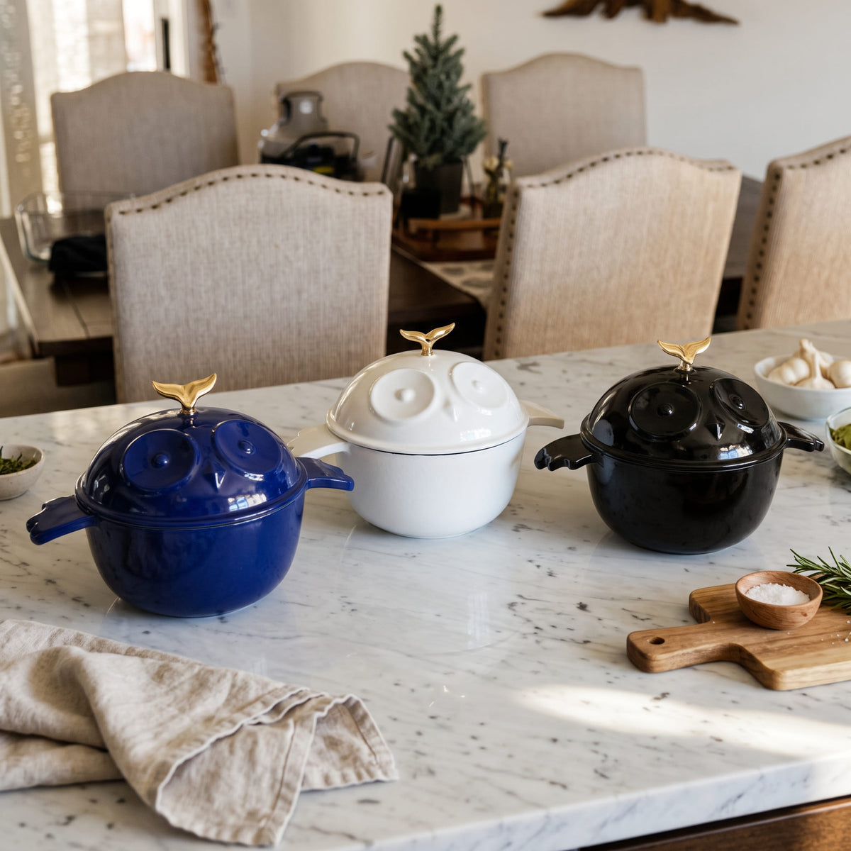 Three decorative enameled lidded pots with gold bird-shaped handles are arranged on a white marble kitchen counter in a dining room setting. The pots are in navy blue, white, and glossy black, with matching enamel cast iron bird-shaped lids and gold finials.