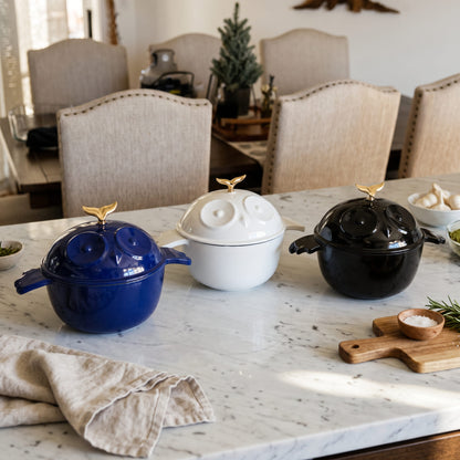 Three decorative enameled lidded pots with gold bird-shaped handles are arranged on a white marble kitchen counter in a dining room setting. The pots are in navy blue, white, and glossy black, with matching enamel cast iron bird-shaped lids and gold finials.
