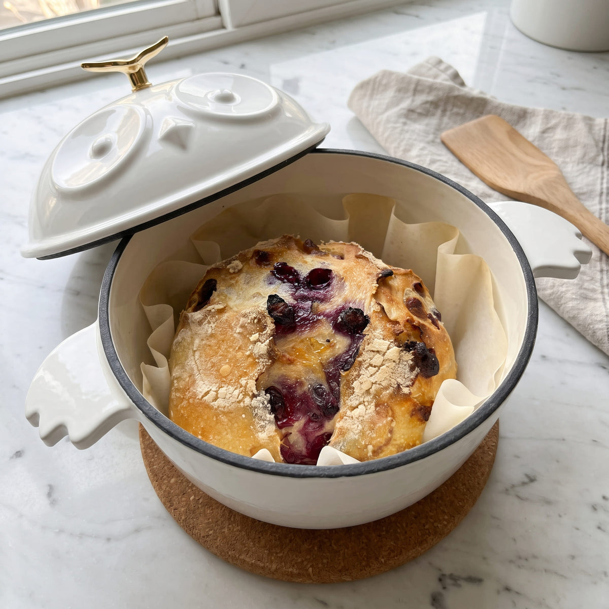 A white enamel cast-iron Dutch oven with a matching lid, shown with its lid off to reveal a baked berry breakfast pie inside. The pot is lined with parchment paper and sits on a cork coaster on a white marble countertop. 1.5 qt