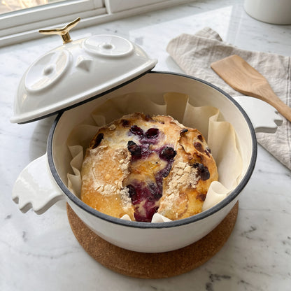 A white enamel cast-iron Dutch oven with a matching lid, shown with its lid off to reveal a baked berry breakfast pie inside. The pot is lined with parchment paper and sits on a cork coaster on a white marble countertop. 1.5 qt