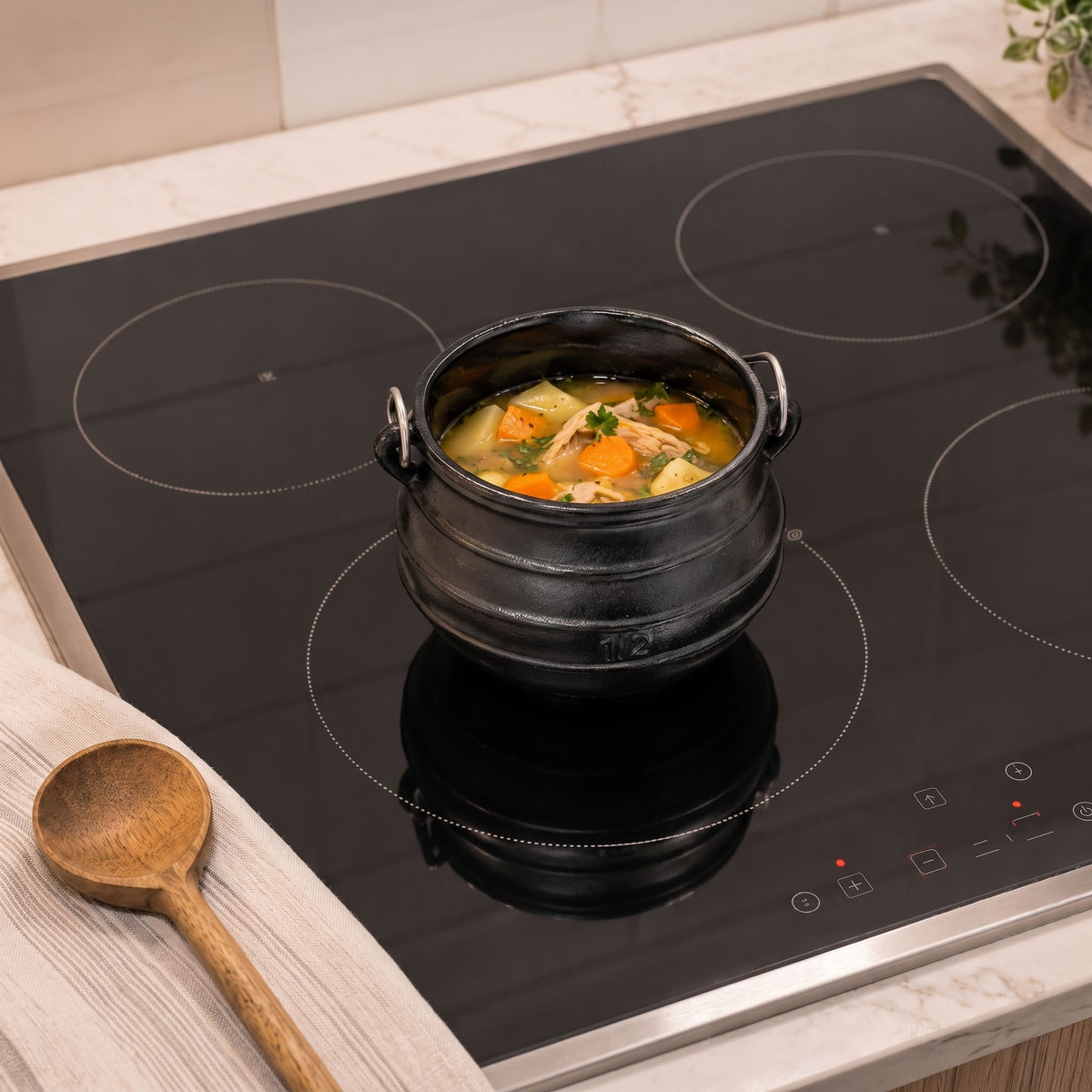 A black ceramic glass cooktop on a light marble countertop, with a black pot of soup cooking on one of its circular heating zones. A wooden spoon rests on a folded white cloth in the foreground. 2 qt cast iron cauldron
