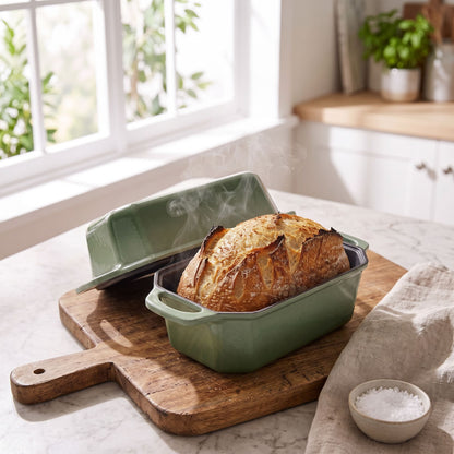 A sage green rectangular baking dish with a lid sits on a wooden cutting board on a kitchen counter, with a freshly baked loaf of bread inside. Steam rises from the bread, and a small bowl of coarse salt is nearby. 8.5x4.5 Inch
