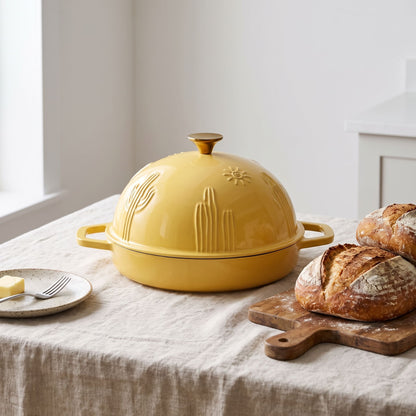 A butter yellow enameled cast iron bread oven with side handles sits on a linen tablecloth in a bright kitchen setting, styled with bread on a wooden board, a small plate with butter, and a fork