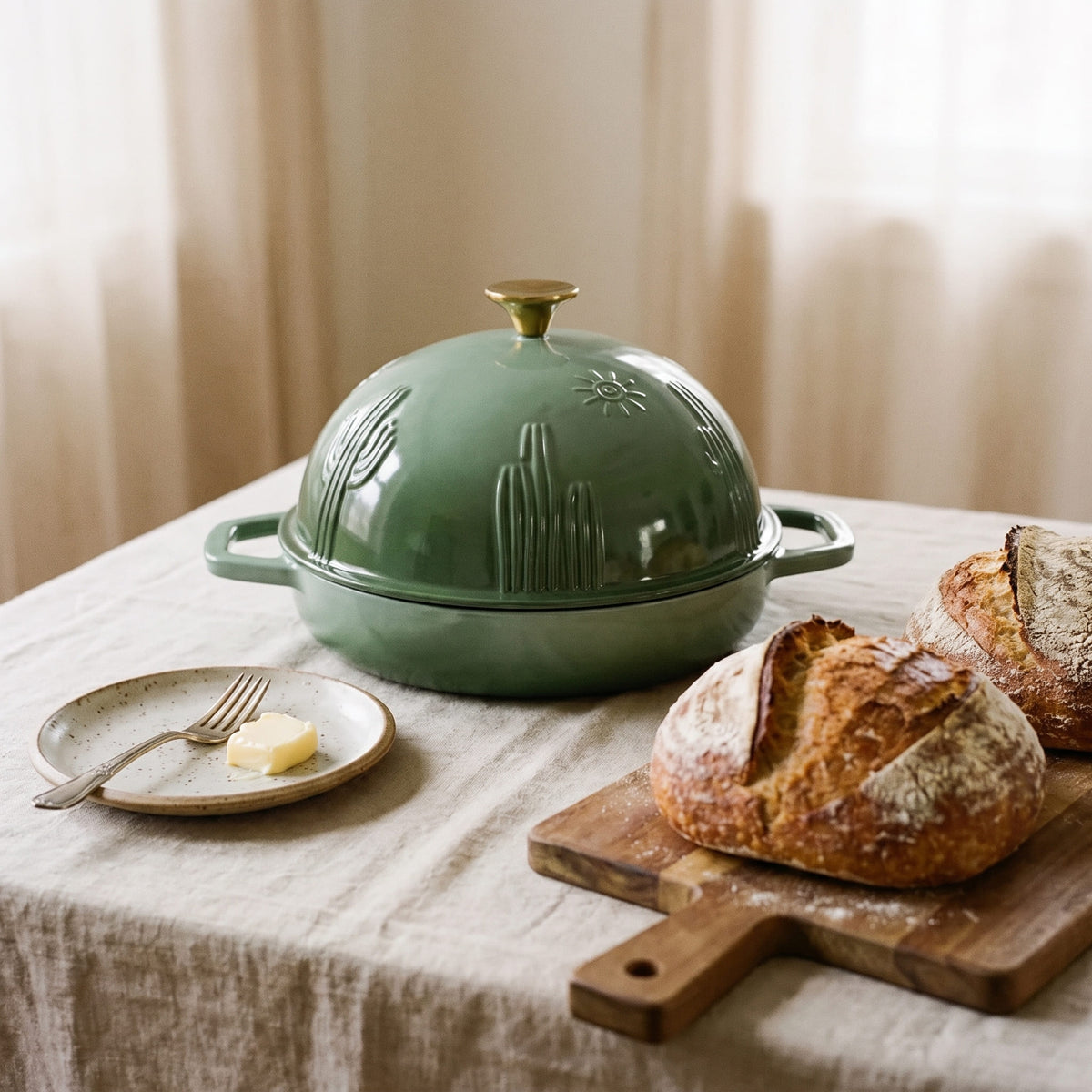 A sage green cast iron bread oven with a gold-toned knob sits on a table covered with a beige linen cloth. A small plate with butter and a fork is beside it, and rustic bread on a wooden board appears in the foreground.