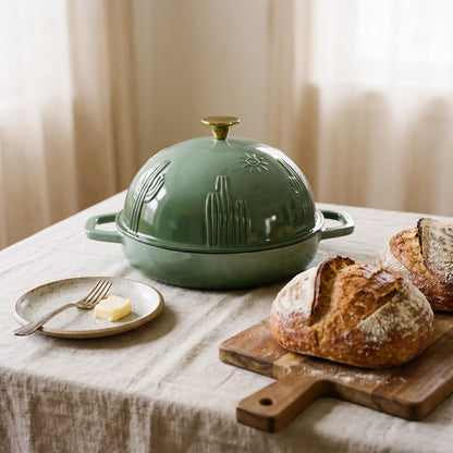 A sage green cast iron bread oven with a gold-toned knob sits on a table covered with a beige linen cloth. A small plate with butter and a fork is beside it, and rustic bread on a wooden board appears in the foreground.