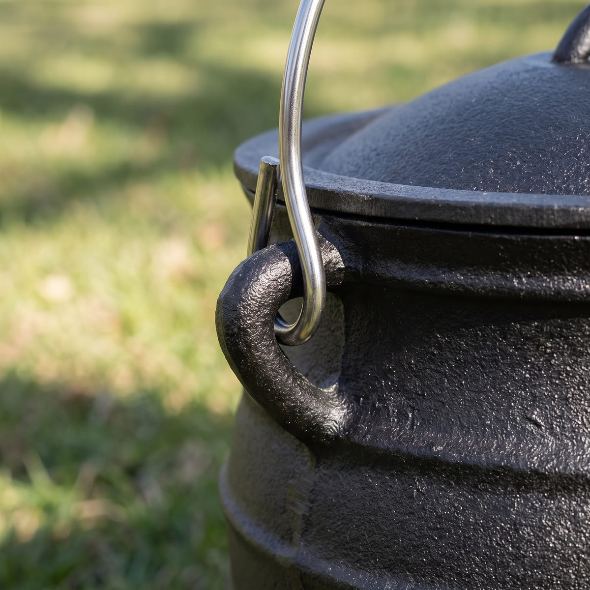 Close-up of a black cast iron potjie with a lid, featuring a curved silver metal handle and side loop, photographed outdoors on a blurred grassy background. 6 qt (Size #2), 8 qt (Size #3), 10 qt (Size #4), 14 qt (Size #6)