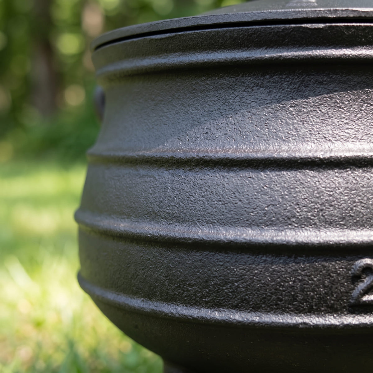 Close-up of a black cast iron pot with horizontal ridges, photographed outdoors with a blurred green grassy background. 6 qt (Size #2), 8 qt (Size #3), 10 qt (Size #4), 14 qt (Size #6)