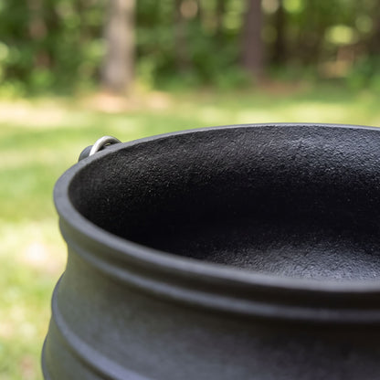 Close-up of a dark cast iron pot potjie rim with a small metal loop handle, photographed outdoors with a blurred green forest background. 6 qt (Size #2), 8 qt (Size #3), 10 qt (Size #4), 14 qt (Size #6)