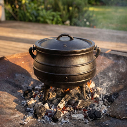 A black cast iron cauldron with a lid sits over glowing embers and coals on an outdoor patio, with greenery blurred in the background. 6 qt (Size #2) cast iron potjie