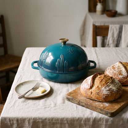 A deep cyan cast iron bread oven with embossed desert cactus designs sits on a linen-covered table next to rustic bread on a wooden board and a small plate with butter and a fork.