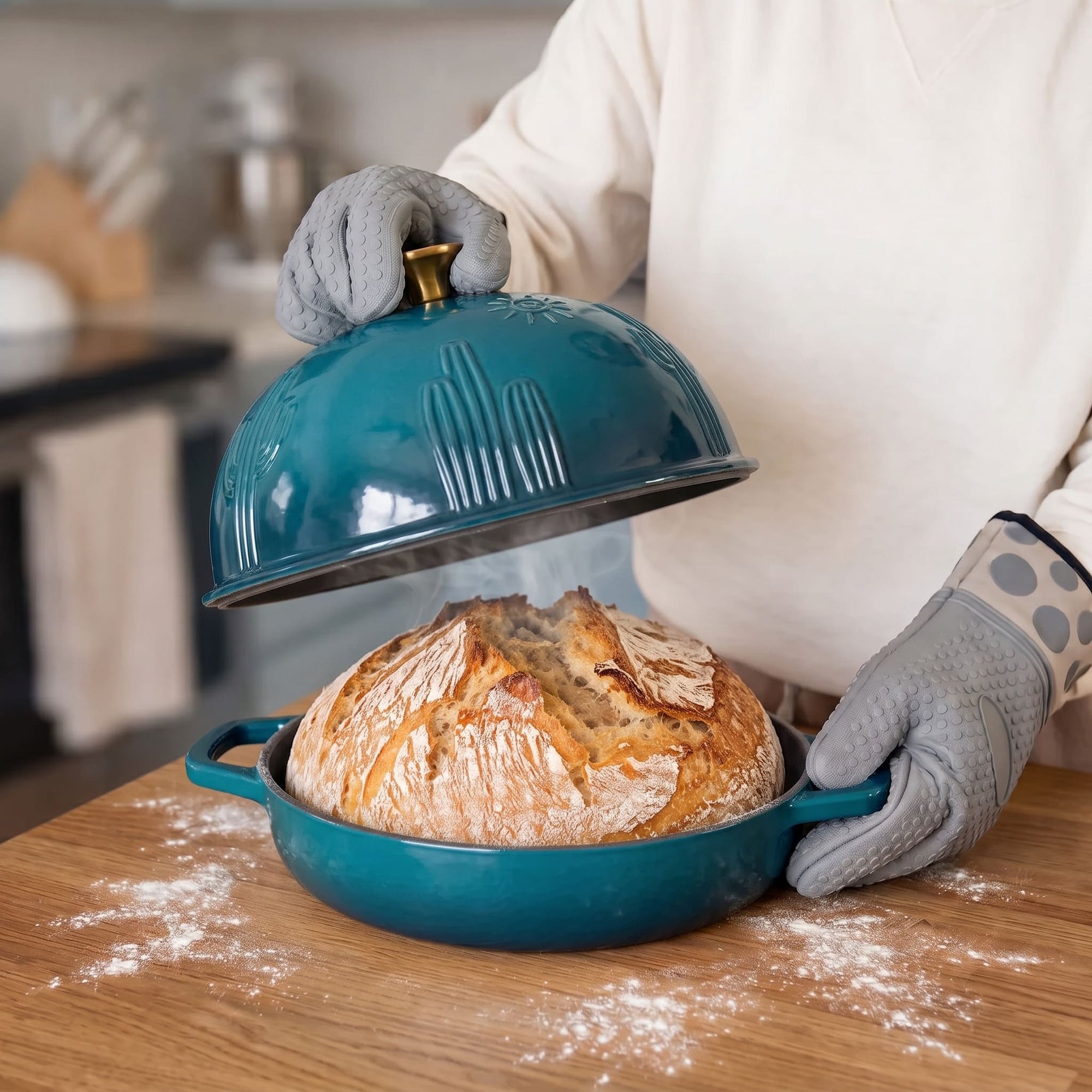 A deep cyan baking dish with a matching lid, shown on a wooden countertop with a loaf of bread rising inside. A person wearing gray oven mitts lifts the lid, with flour scattered on the counter in a kitchen setting.