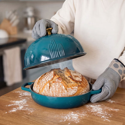 A deep cyan baking dish with a matching lid, shown on a wooden countertop with a loaf of bread rising inside. A person wearing gray oven mitts lifts the lid, with flour scattered on the counter in a kitchen setting.