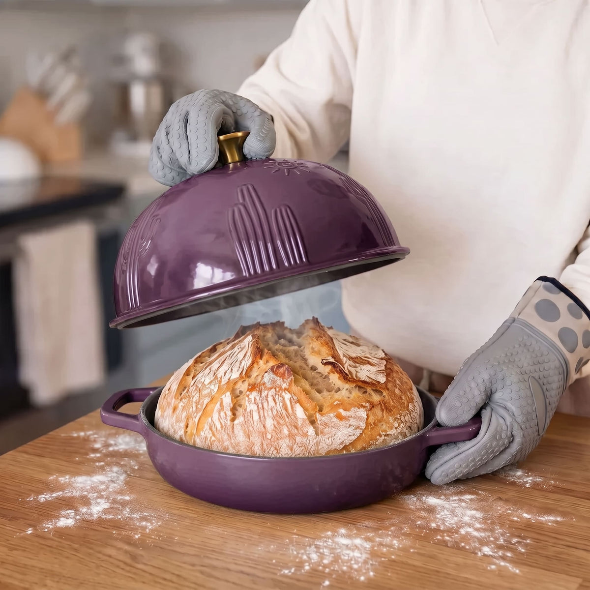 A person wearing gray oven mitts lifts a plum purple dome lid off a round purple baking dish containing a freshly baked sourdough bread loaf, set on a wooden table with flour dusting the surface.