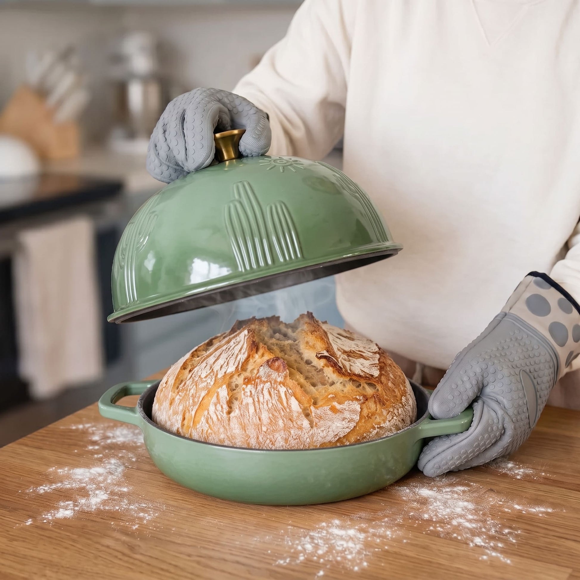 A person wearing gray oven mitts lifts the lid off a sage green baking dish containing a freshly baked sourdough bread loaf, set on a wooden table with flour scattered around.