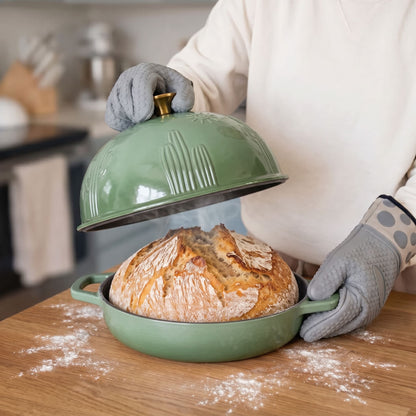 A person wearing gray oven mitts lifts the lid off a sage green baking dish containing a freshly baked sourdough bread loaf, set on a wooden table with flour scattered around.