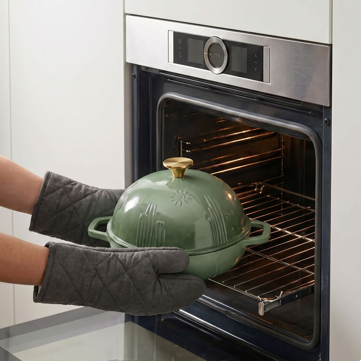 A sage green bread oven with a gold knob is being pulled from an oven by two hands wearing gray quilted oven mitts. The oven is stainless steel with a dark glass door.