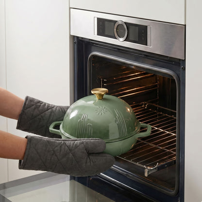 A sage green bread oven with a gold knob is being pulled from an oven by two hands wearing gray quilted oven mitts. The oven is stainless steel with a dark glass door.