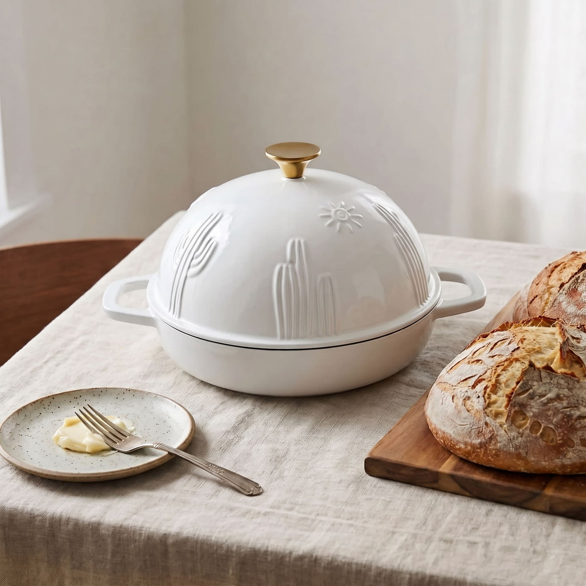 A pearl white cast iron lidded bread pan with a gold-toned knob and embossed cactus and sun motifs, set on a linen tablecloth beside a plate with butter and fork and a loaf of bread on a wooden board.