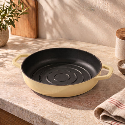 A round cast iron with a butter yellow exterior and a black interior, shown on a beige stone countertop in a kitchen setting.