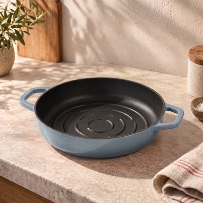 A round cast iron bread pan with a misty blue exterior and a black interior, resting on a beige stone countertop in a kitchen setting.