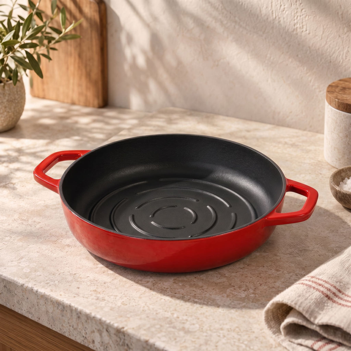 A cherry red enameled cast iron bread pan with a black interior sits on a beige stone countertop in a kitchen setting with natural sunlight and shadows.
