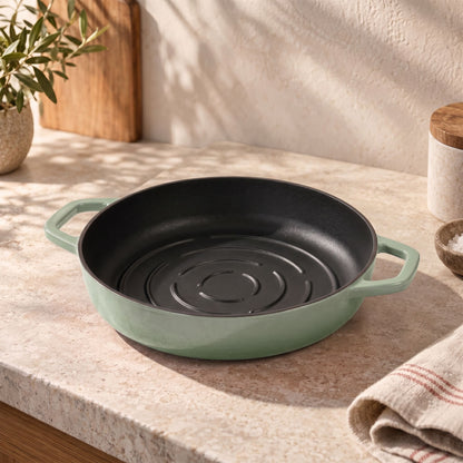 A round cast iron bread pan with a sage green exterior and a black non-stick interior sits on a beige stone countertop in a kitchen setting.