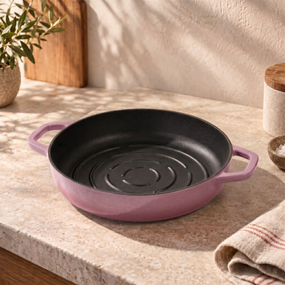 A round cast iron bread pan with a rose pink exterior and a black non-stick interior, styled on a beige stone countertop with a potted plant and kitchen items in the background.