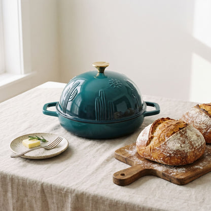 Teal enamel bread oven with domed lid, holding a round loaf on a table.