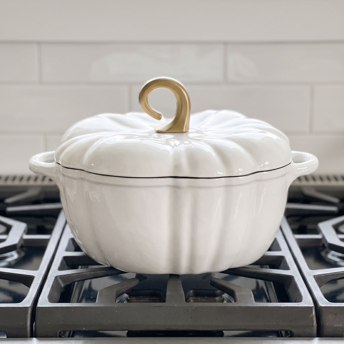 A white enameled cast iron Dutch oven with a lid designed to look like a pumpkin, featuring a gold-colored loop handle and a thin black rim, sitting on a gas stovetop. pearl white