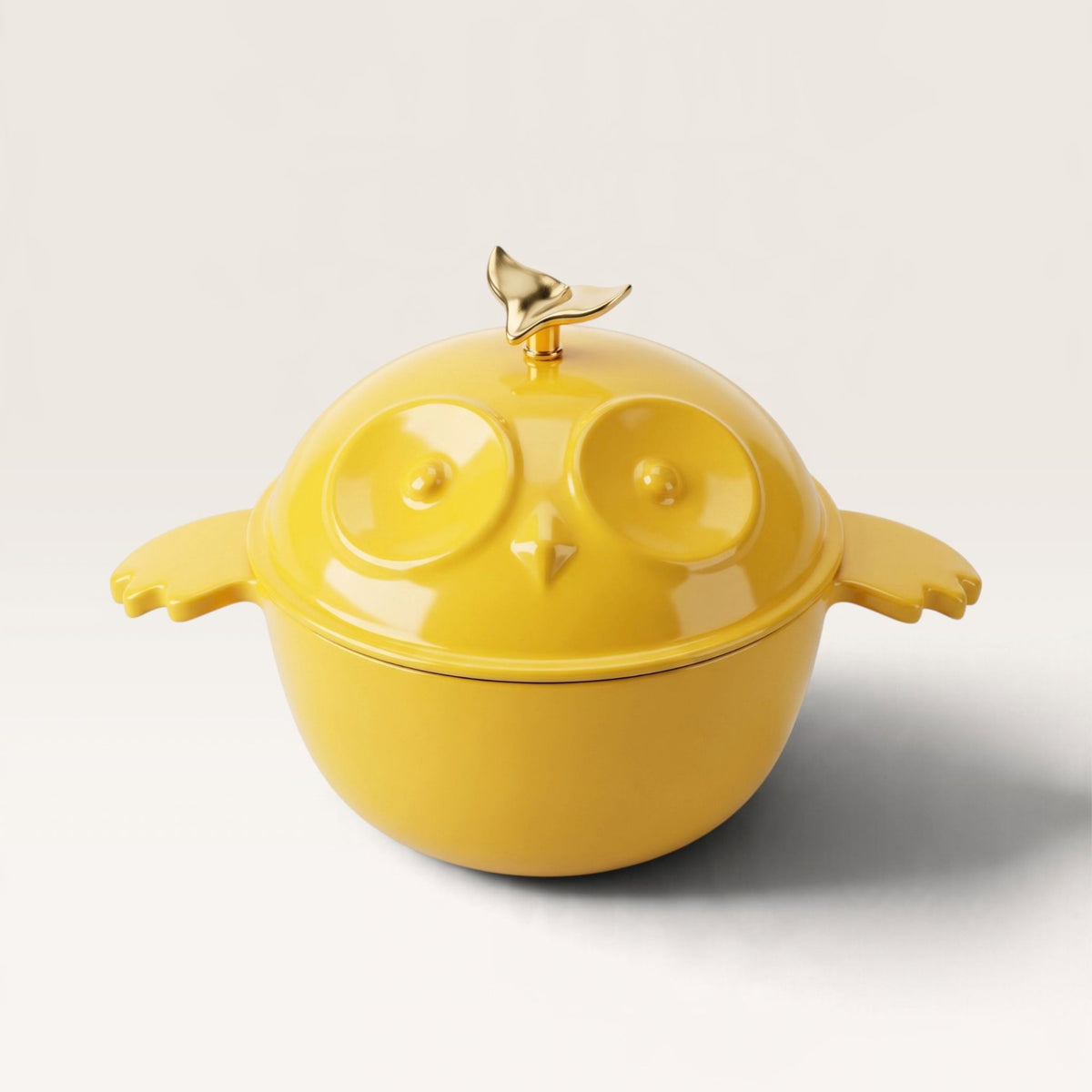 A bright yellow owl-shaped lidded dutch ovenwith winged side handles and a metallic gold bird topper on the lid, photographed on a clean light gray studio background.