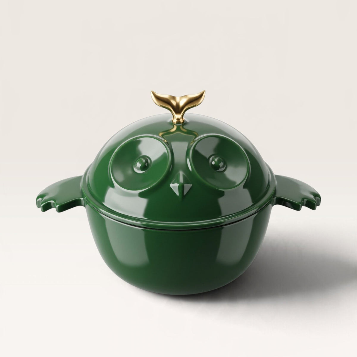 A glossy dark green cast iron dutch oven shaped like an owl, featuring raised owl eyes, small wing-like side handles, and a metallic gold bird-tooth style lid handle, photographed on a clean light gray studio background.