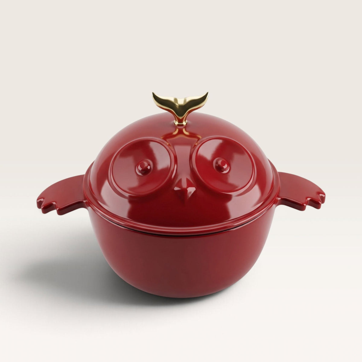 A glossy red owl-shaped lidded pot dutch oven with side handles and a gold bird-toy-shaped lid knob, photographed on a clean light gray studio background.