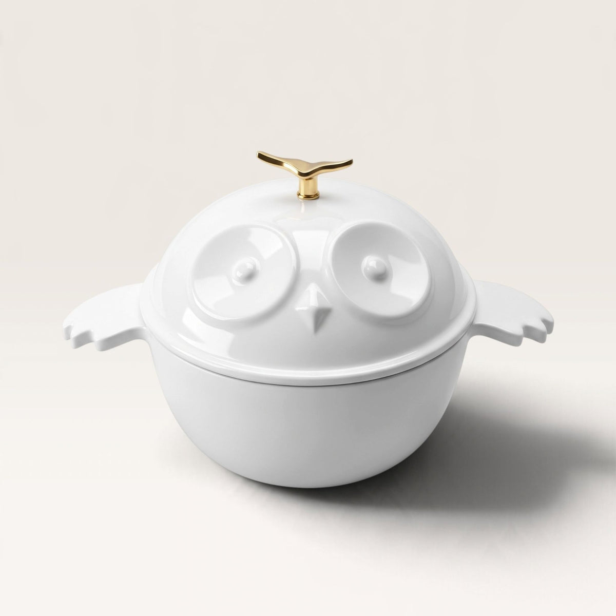 A white enameled cast iron owl-shaped lidded dutch oven with sculpted wings on both sides and a small gold handle on top, photographed on a clean light beige studio background.