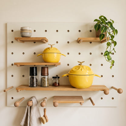 A wall-mounted pegboard kitchen organizer with light wood shelves and hooks, styled with two bright yellow lidded pots, spice jars, stacked bowls, a hanging towel, a scrubber, and a trailing plant against a wall.