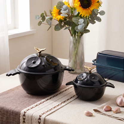 Two glossy black ceramic stock pots with lids and gold-toned bird-shaped handles, styled on a table runner with sunflowers, garlic, and a loaf pan in the background.