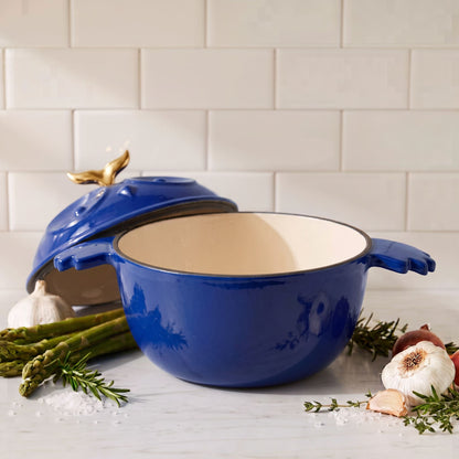 A cobalt blue enameled cast iron Dutch oven with a lid featuring a gold leaf handle, set on a marble countertop with garlic, asparagus, and rosemary against a subway tile backsplash.