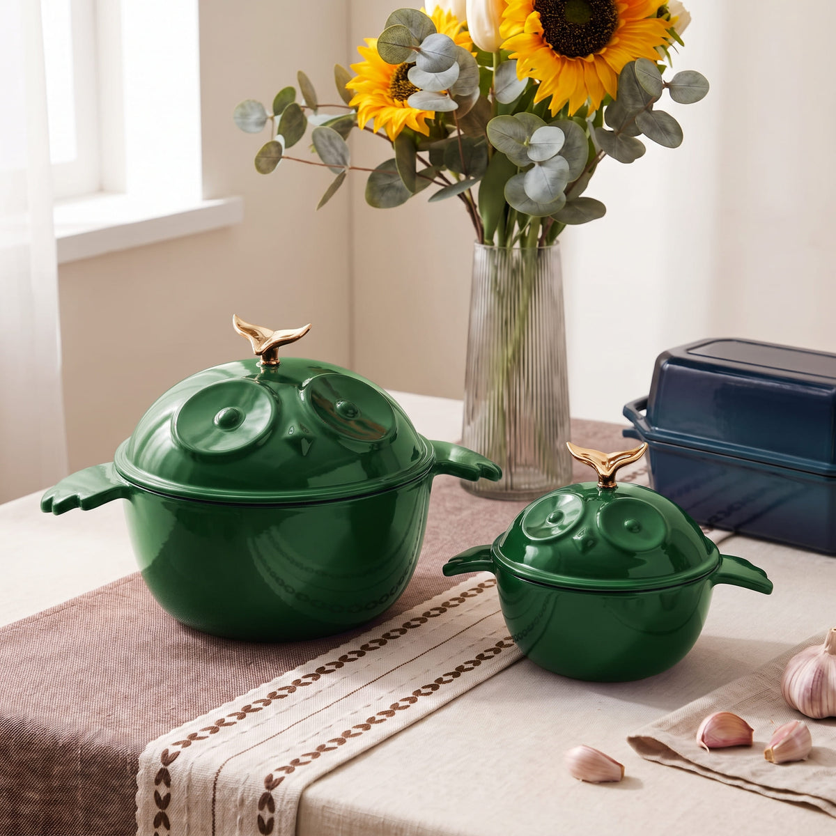 Two green enameled lidded pots with decorative knob handles and lids, styled on a table with a floral vase, sunflowers, garlic, and a loaf pan in a bright indoor setting.