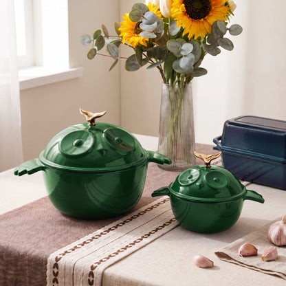 Two green enameled lidded pots with decorative knob handles and lids, styled on a table with a floral vase, sunflowers, garlic, and a loaf pan in a bright indoor setting.
