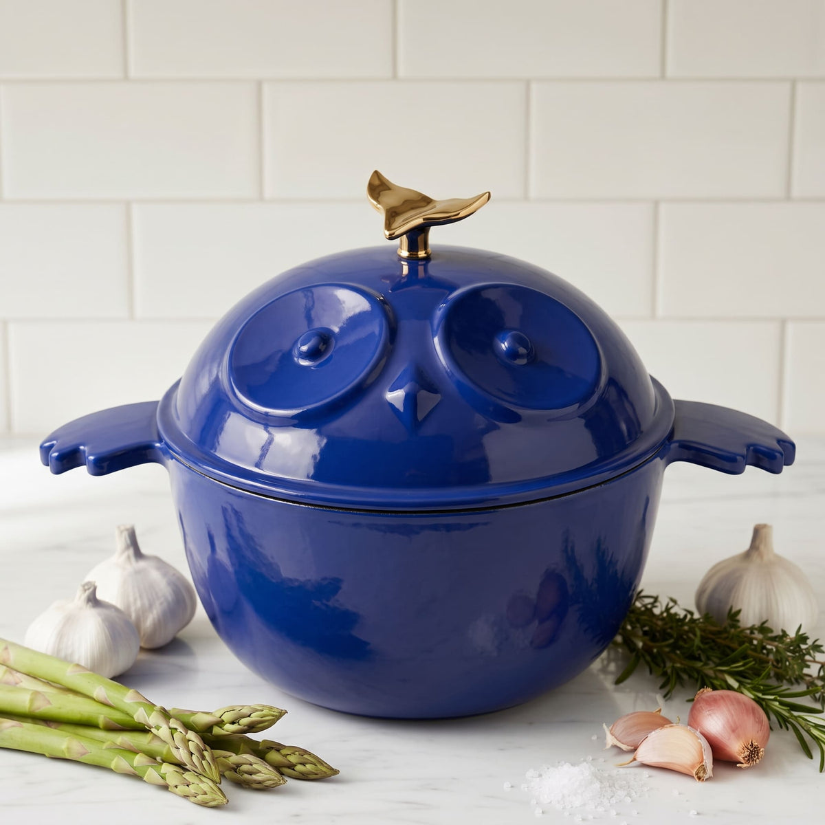 A glossy cobalt blue enamel pot with a matching lid shaped like an owl, featuring a small gold bird finial on top. The pot sits on a marble countertop surrounded by cooking materials, including garlic, asparagus, shallots, rosemary, and sea salt, with a subway tile backsplash in the background.