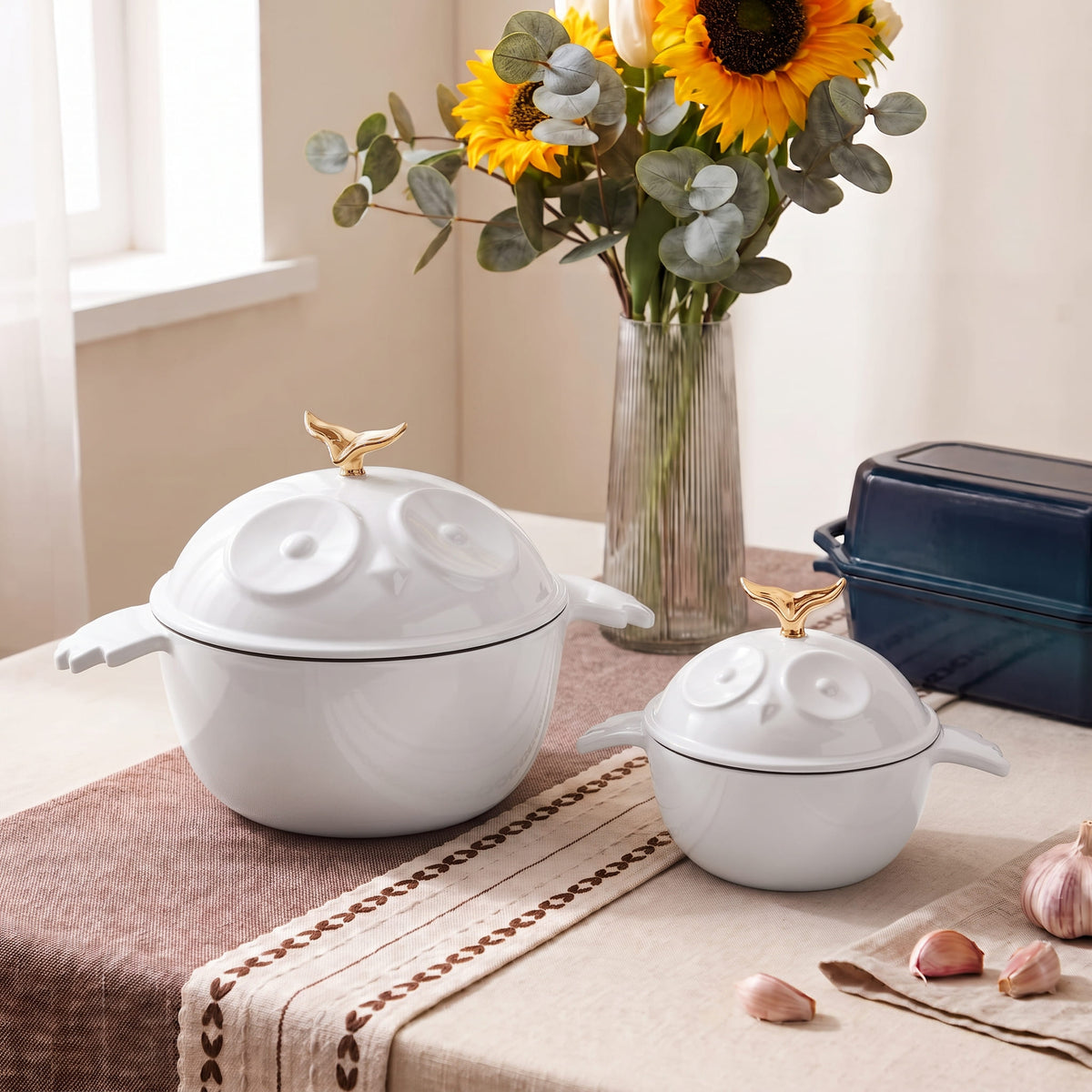 Two white cast iron owl-shaped lidded pots with gold bird toppers sit on a table runner in a styled kitchen setting, with a vase of sunflowers and garlic in the background.
