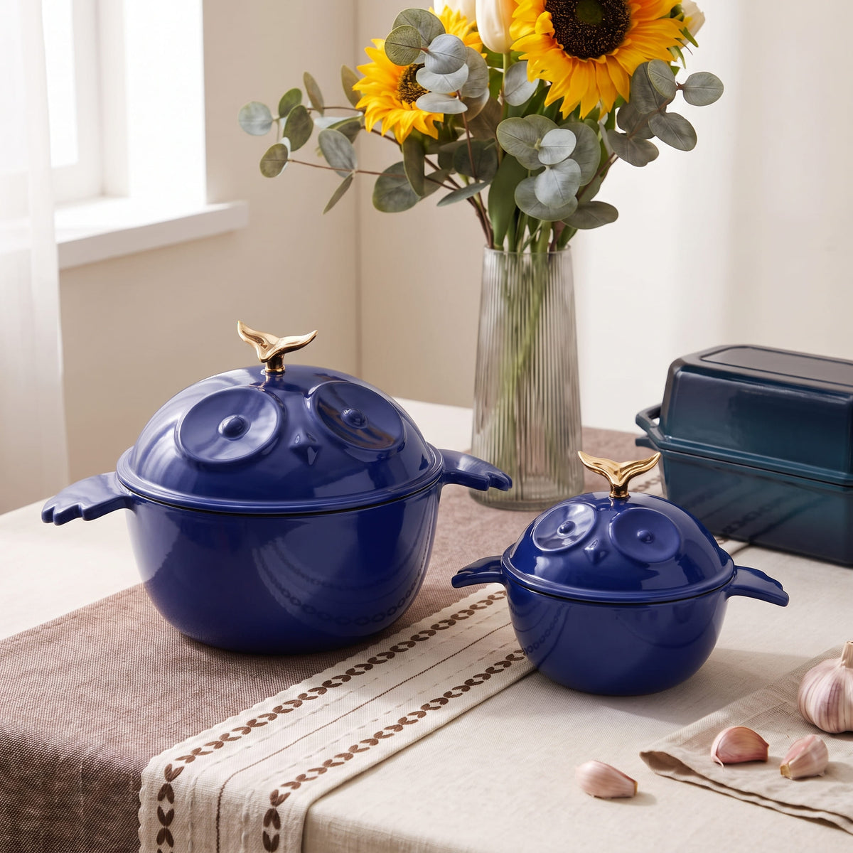 Two dark blue enamel lidded pots with gold bird-shaped handles sit on a beige table runner in a softly lit kitchen setting. A vase of sunflowers and eucalyptus, garlic, and a dark rectangular container are arranged around them.