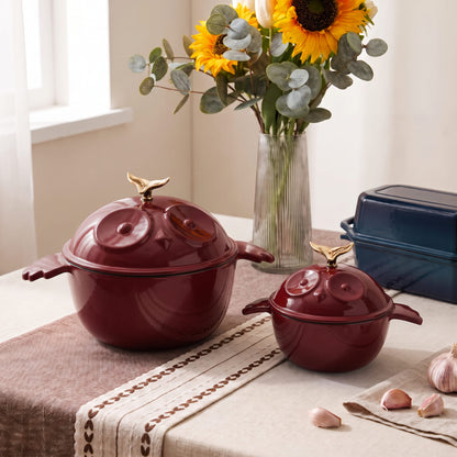 Two glossy deep red enameled lidded pots with gold bird-shaped lid handles sit on a beige table runner. A vase of sunflowers and eucalyptus, garlic cloves, and a liddedloaf pan are arranged around them in a warm kitchen setting.