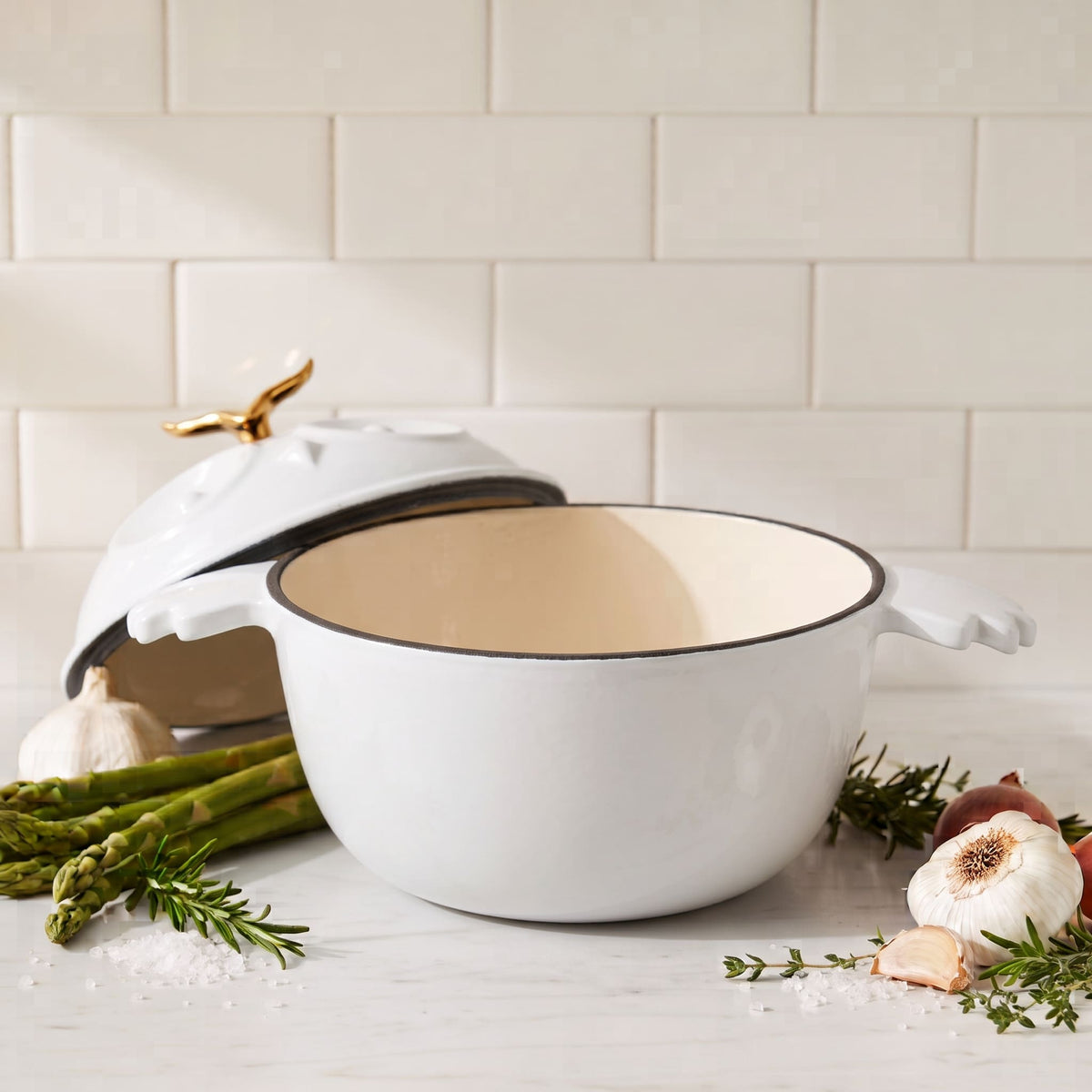 A white enameled cast iron Dutch oven with a lid, styled on a white marble countertop with garlic, asparagus, and rosemary against a white subway tile backsplash.
