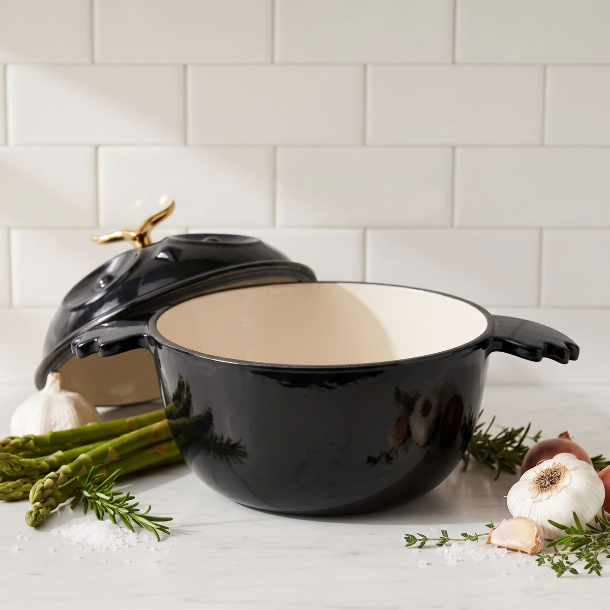 A black enameled cast iron Dutch oven with a cream interior and a gold-toned lid handle, styled on a marble countertop with garlic, asparagus, rosemary, and sea salt against a subway tile backsplash.