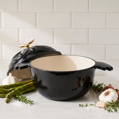 A black enameled cast iron Dutch oven with a cream interior and a gold-toned lid handle, styled on a marble countertop with garlic, asparagus, rosemary, and sea salt against a subway tile backsplash.