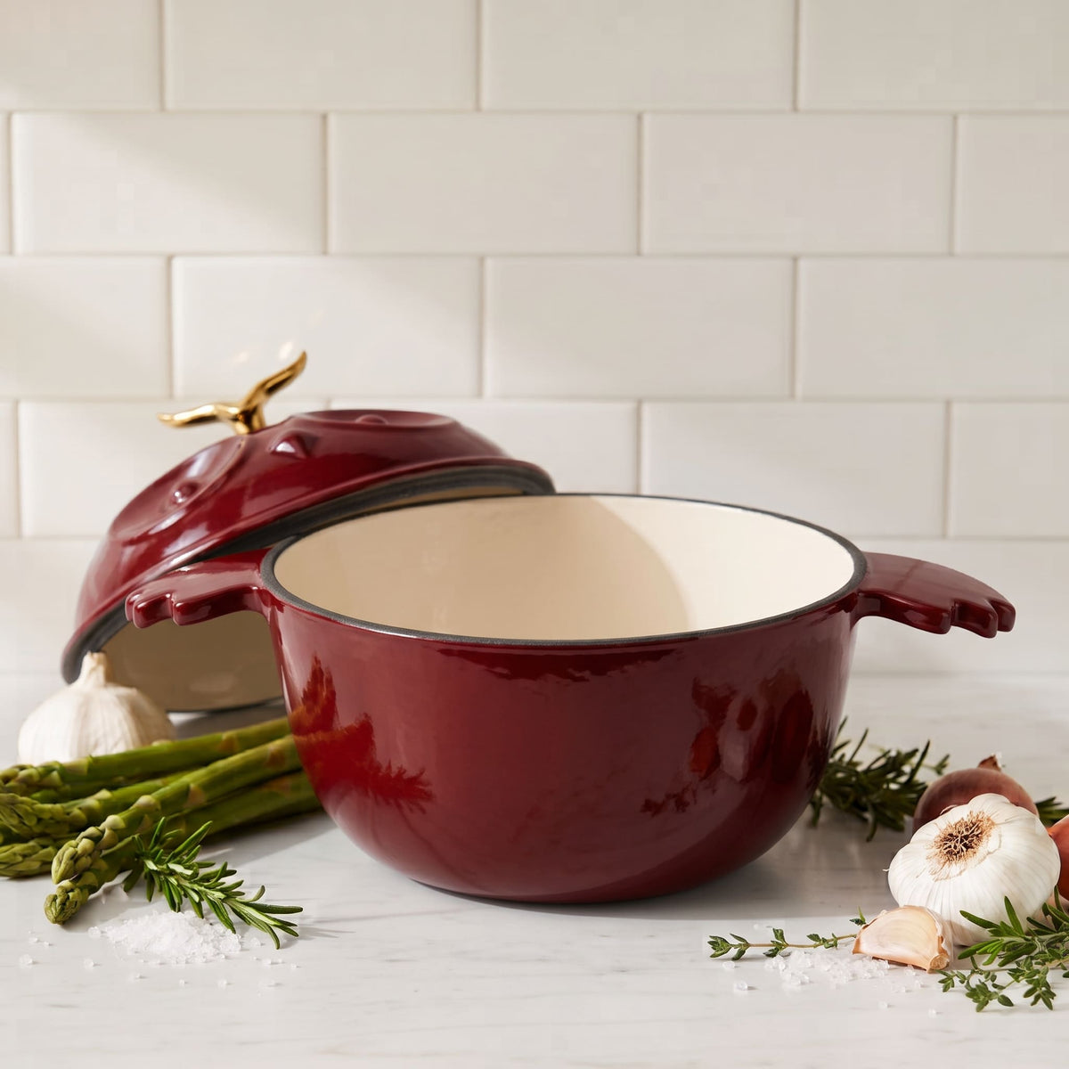 A deep red enameled cast iron pot with a cream-colored interior and gold-toned lid handle, styled on a marble countertop with garlic, asparagus, and rosemary against a subway tile backsplash.