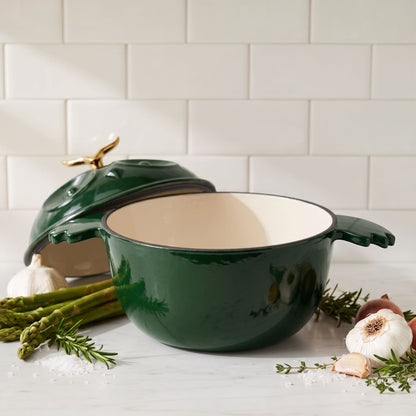 A dark green enameled cast iron Dutch oven with a matching lid and gold-toned handle knob, styled on a marble countertop with garlic, asparagus, and rosemary against a subway tile backsplash.