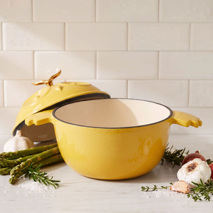 A yellow enameled cast iron Dutch oven with a lid, styled on a marble countertop with garlic, asparagus, and rosemary against a subway tile backsplash.