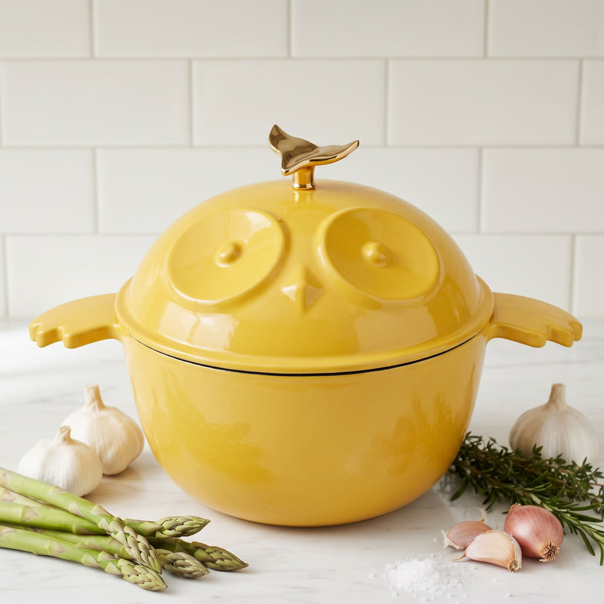 A bright yellow owl-shaped lidded pot with bird-shaped handles and a gold bird topper, styled on a marble surface with garlic, asparagus, rosemary, and shallots against a subway tile background.
